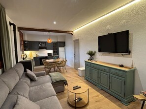 Ein modernes Wohnzimmer mit grauem Sofa, einer Essecke mit rundem Tisch und Stühlen, einer Küche mit dunklen Schränken und einem an der Wand befestigten Fernseher.