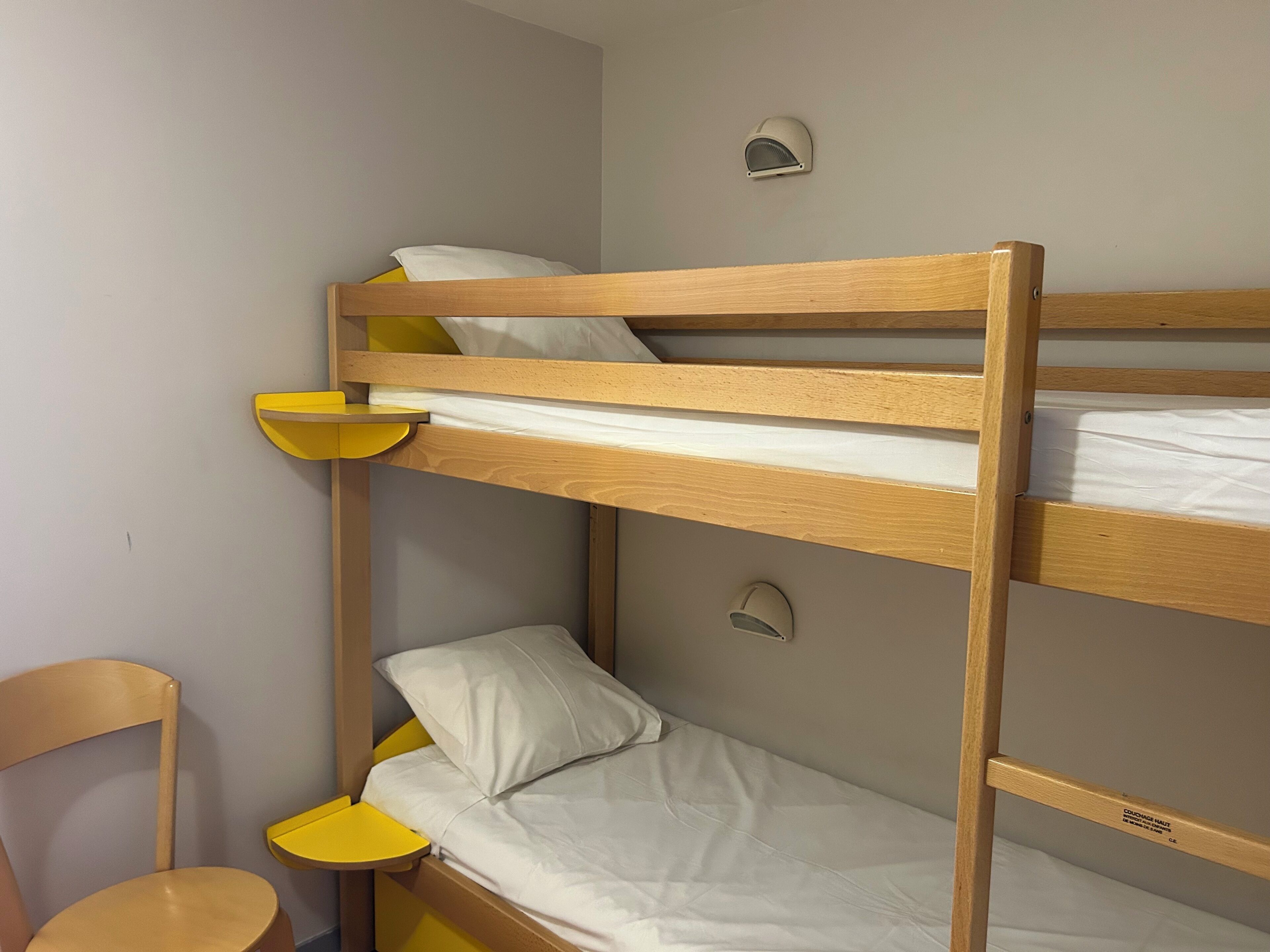 A bunk bed with a yellow side table and a chair, a small wall-mounted light, and a visible label on the bed frame.