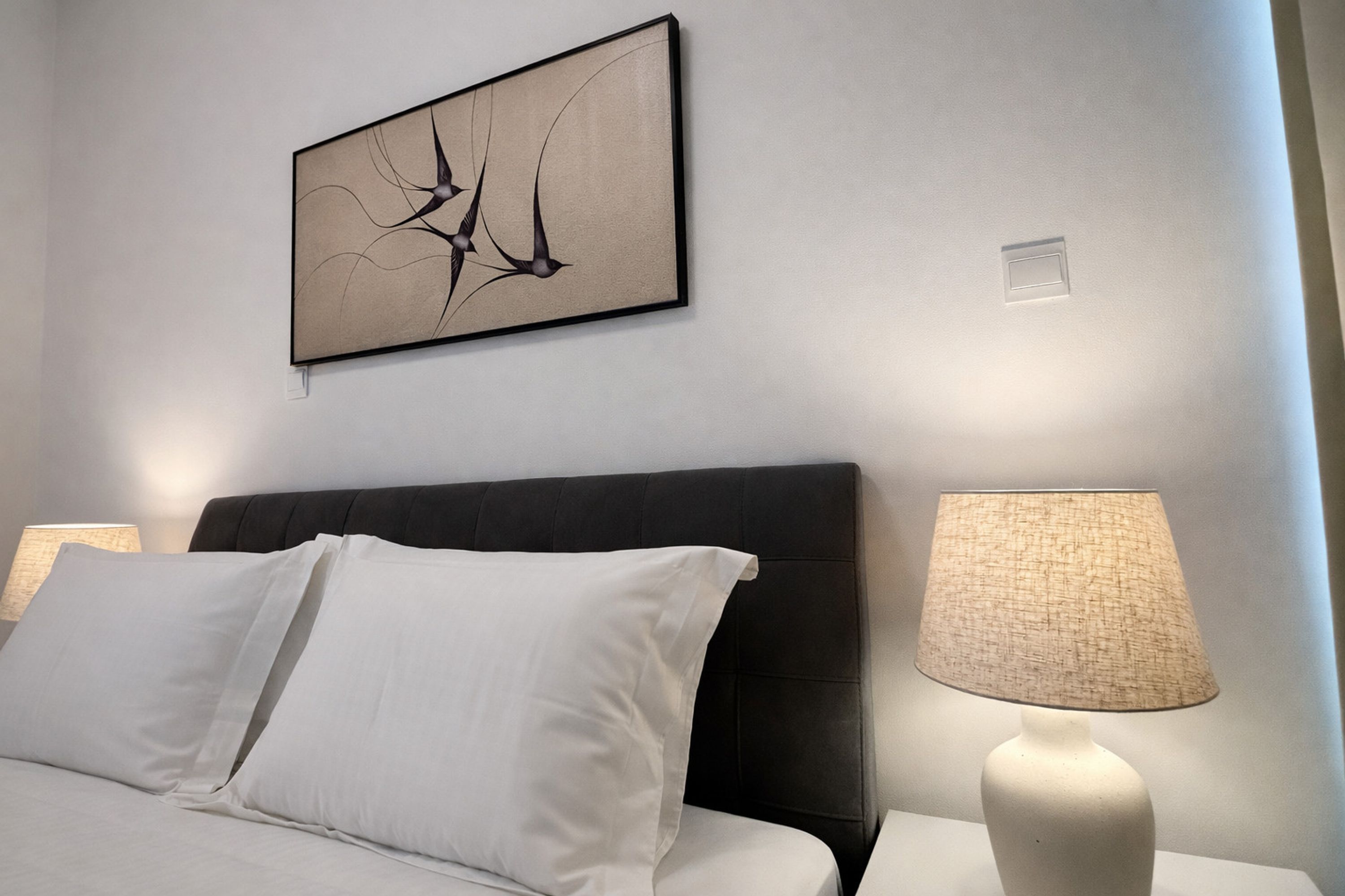 A neatly made bed with white linens, a dark headboard, a wall-mounted abstract art piece, and a bedside lamp.
