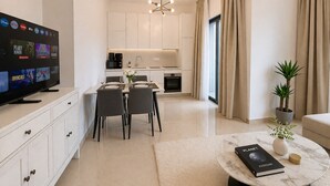 A modern living room with a flat-screen TV, a dining area with a table and chairs, a kitchen with white cabinets, and a round coffee table with a book and a plant. - Oni Residence Apartment (Limassol)