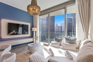 Ein modernes Hotelzimmer mit großem Fernseher, rundem Couchtisch, einer Couch mit vielen Kissen und Blick auf die Stadt durch große Fenster.