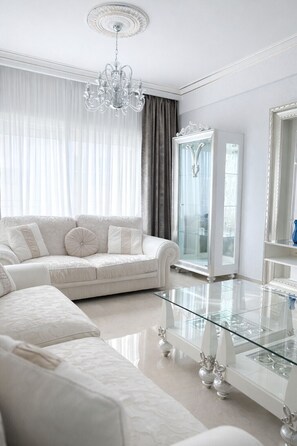 A modern living room with a glass coffee table, a white sofa, a chandelier, and a glass cabinet. - Gulf Beach Apartment (Limassol)
