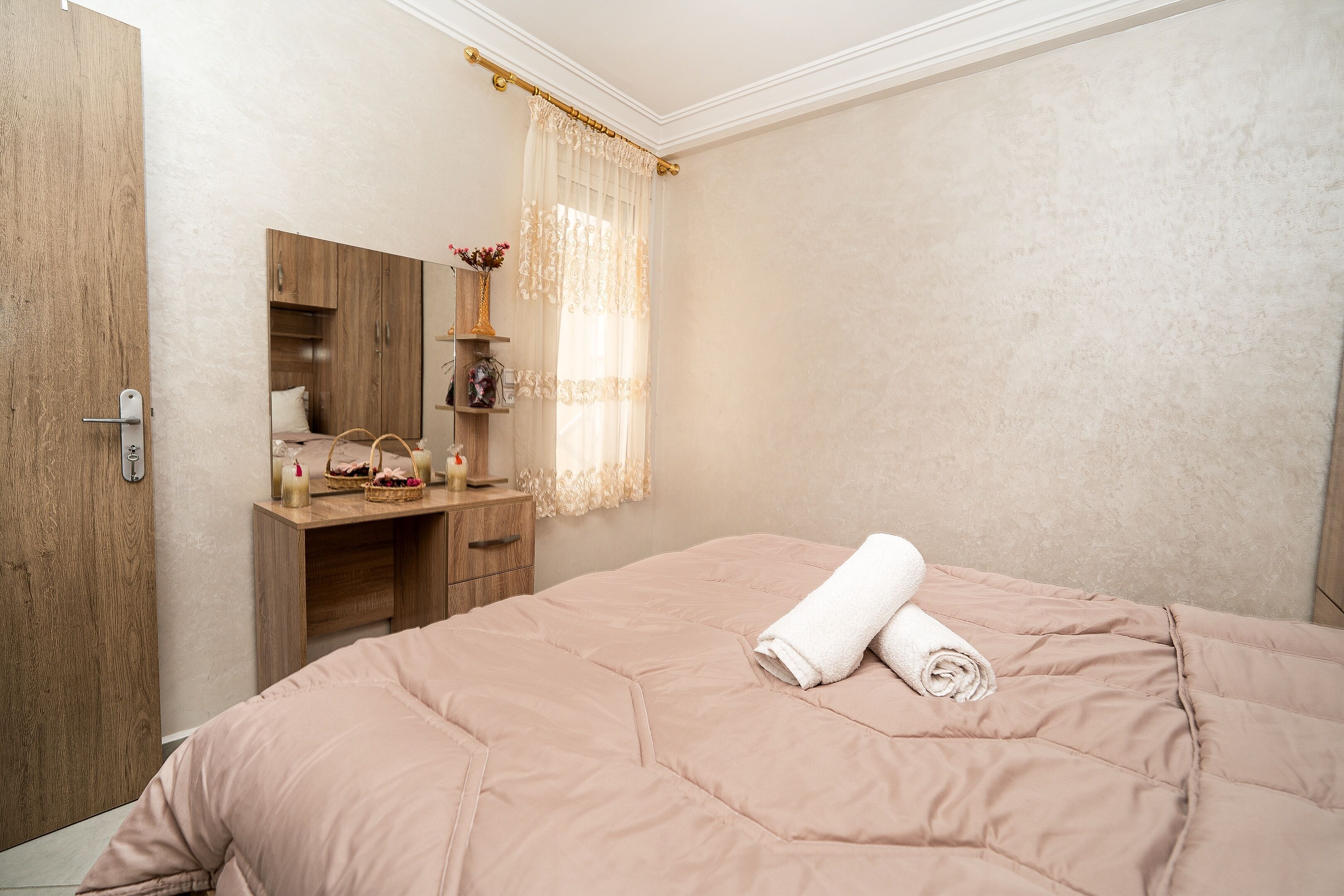 Une chambre à coucher avec un grand lit, une commode en bois, un miroir et une fenêtre ornée de rideaux légers.