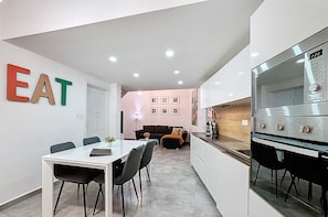 Una cocina moderna con horno empotrado, una zona de comedor con mesa y sillas blancas, y una sala de estar con sofá y mesa de centro.