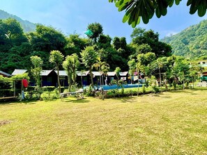 A grassy area with several small, single-story buildings with sloped roofs, surrounded by dense greenery and hills in the background. - Hills Queen Resort & Camping (Mohan Chatti)