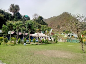 A residential area with houses, a grassy field, and a hill in the background. - Hills Queen Resort & Camping (Mohan Chatti)
