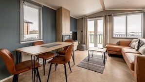 A modern living room with a sofa, dining table, and chairs, a kitchen area, and large windows with curtains.