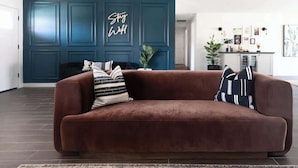 Un sofá marrón con almohadones estampados en una habitación con paredes color turquesa y un cartel que dice 'Stay Well'.