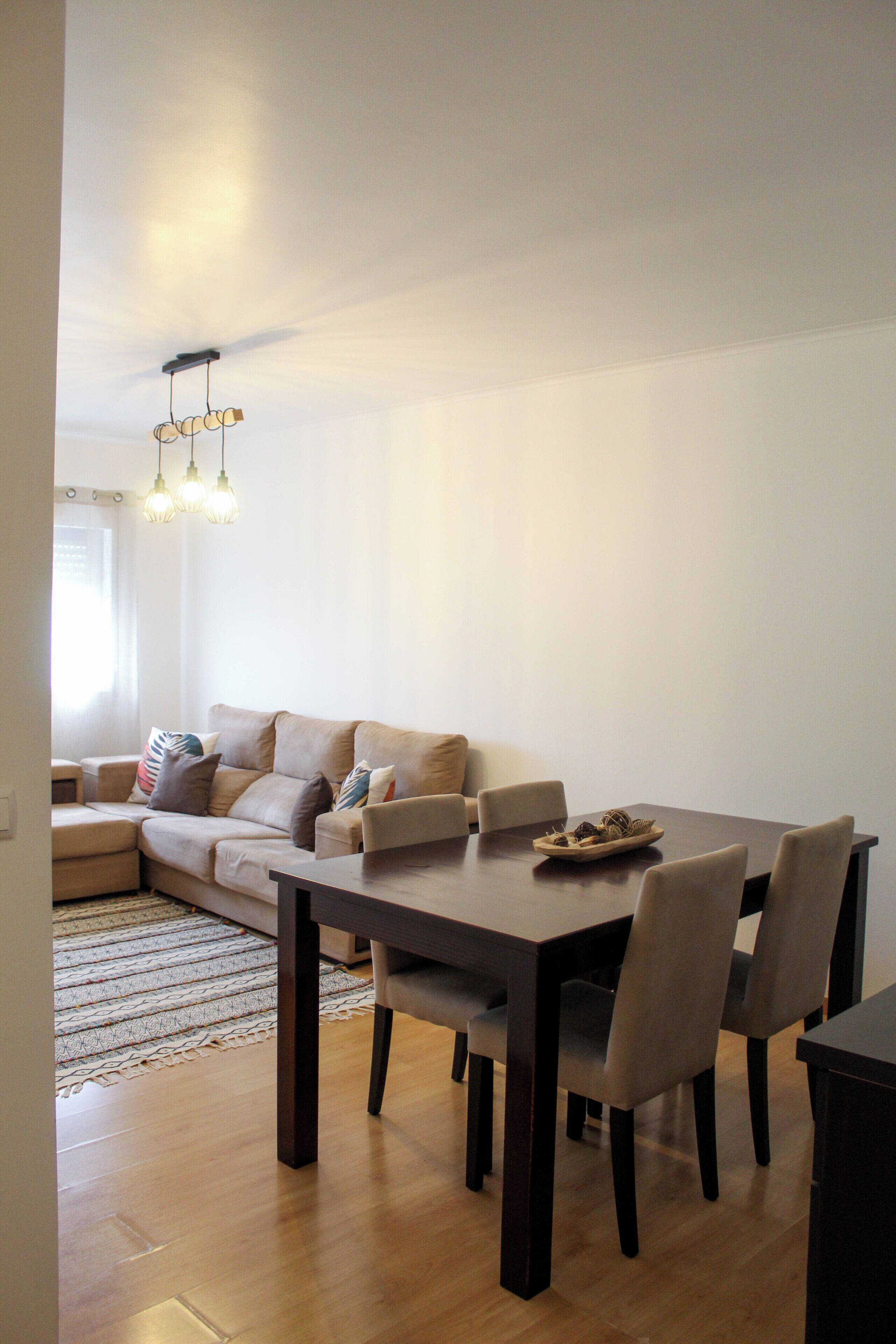 A dining area with a wooden table, four chairs, a sofa, and a hanging light fixture.