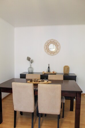 Un coin repas avec une table en bois foncé, des chaises recouvertes d’un tissu beige, un vase orné de feuilles décoratives, un petit miroir rond et un plateau en bois contenant divers objets.