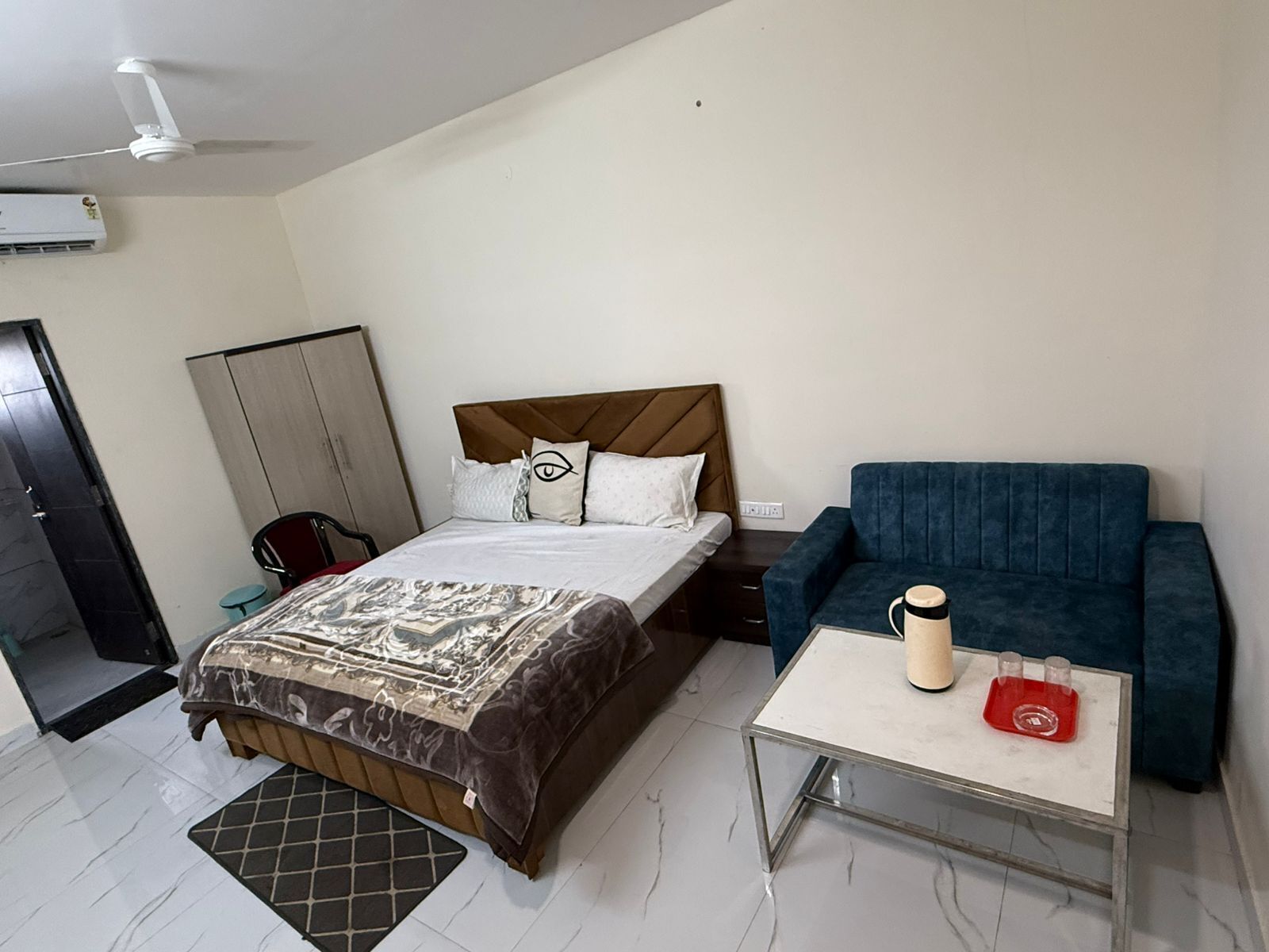 A bedroom with a bed, a blue sofa, a coffee table, a ceiling fan, and an air conditioning unit.