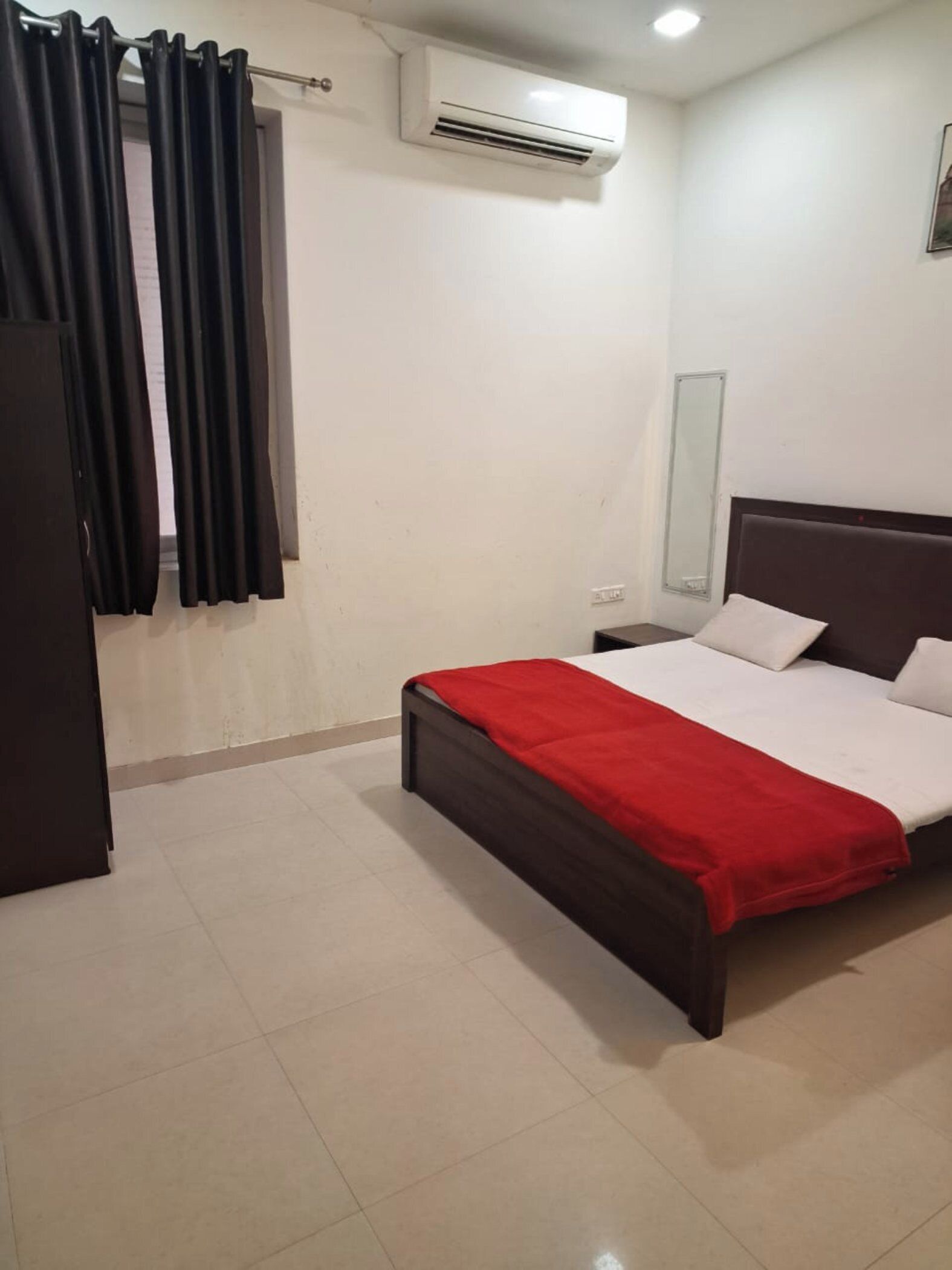 A hotel room with a bed, a red blanket, a dark headboard, a white wall, an air conditioning unit, and a window with black curtains.