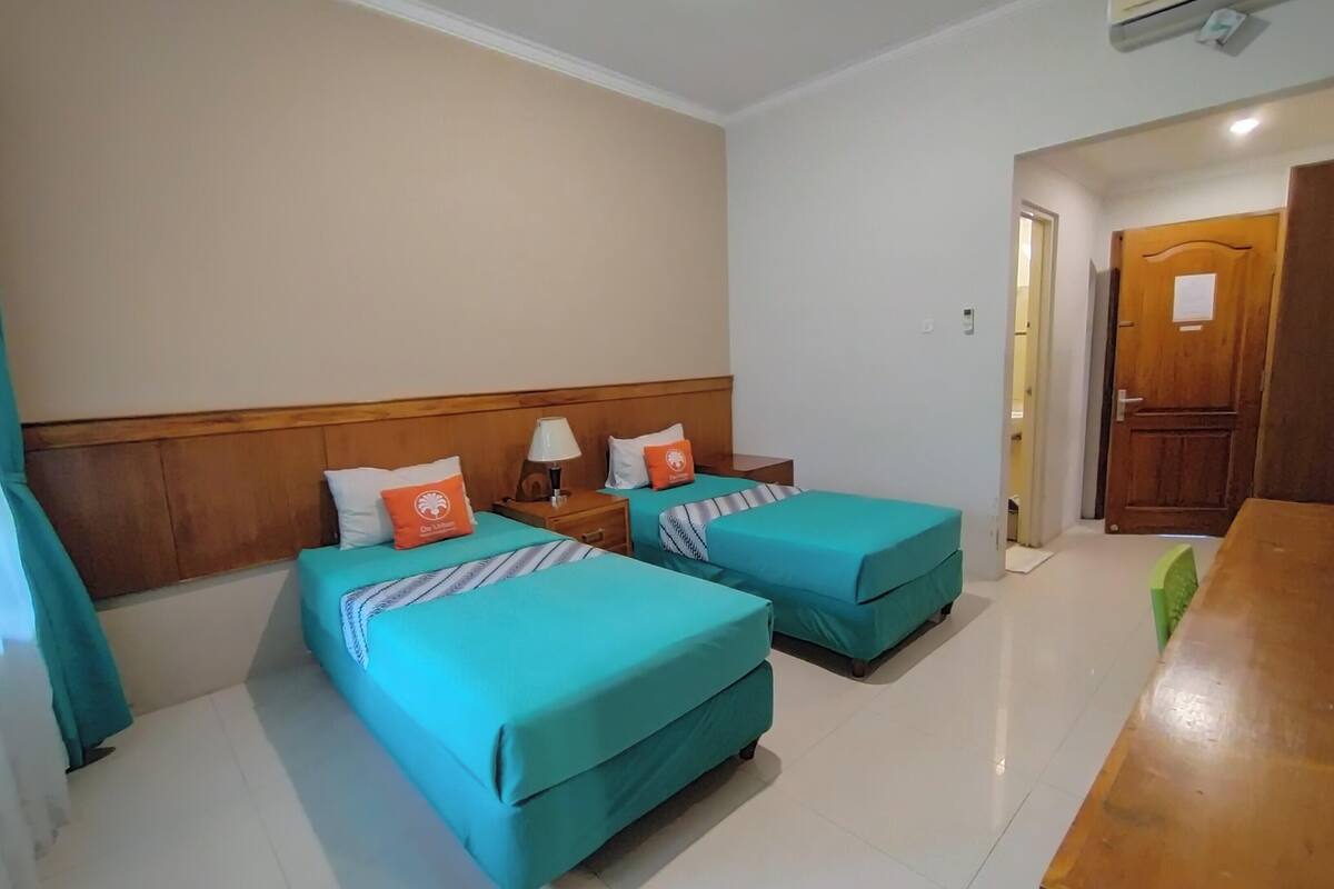Habitación de hotel con dos camas, cabecero de madera, mesita de noche con lámpara y una mesa de madera.