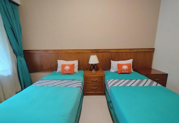 A hotel room with two beds, a wooden headboard, a nightstand with a lamp, and two orange pillows with a logo. - HOTEL URBAN LATIFA YOGYAKARTA (Depok)