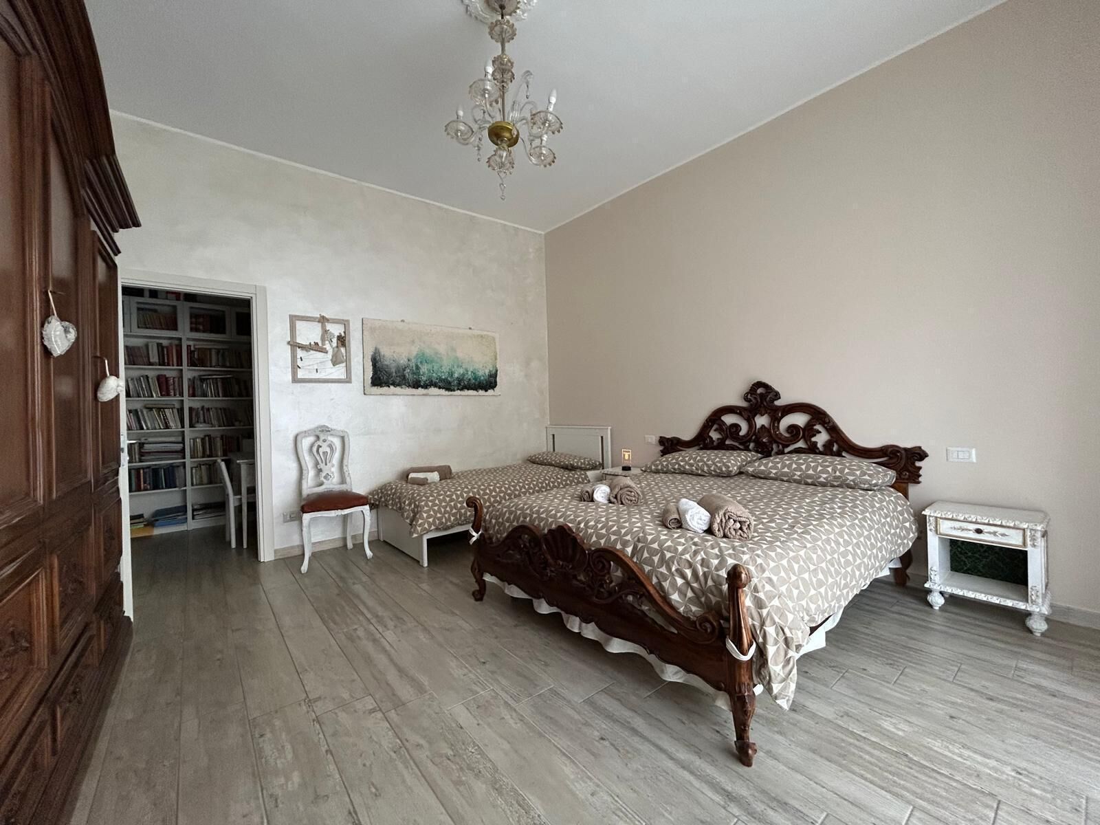 A bedroom with two beds, a wooden floor, a chandelier, a bookshelf, and a painting.