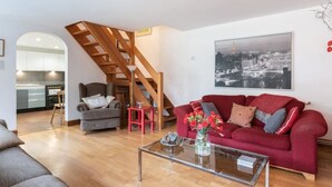 Una sala de estar con un sofá rojo, una mesa de centro de vidrio, un sillón gris, una escalera de madera y una cocina al fondo.