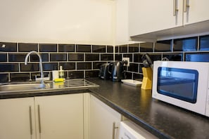 Una cocina con un lavamanos, protector contra salpicaduras de azulejos negros, un hervidor, un microondas y un bloque de cuchillos.