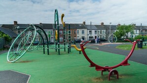 A playground with various play structures, a green rubberized surface, and residential buildings in the background. - Praise Suite | Stylish Apartment • Parking • Wi-Fi (Barry)