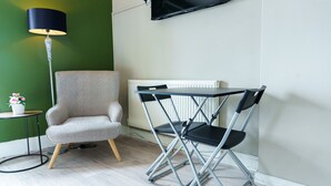 A room with a grey armchair, a black folding chair, a floor lamp, and a TV mounted on the wall. - Praise Suite | Stylish Apartment • Parking • Wi-Fi (Barry)