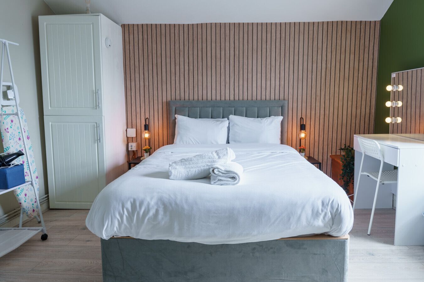 Un lit bien fait, avec une tête de lit grise, une table de chevet avec une lampe et un miroir avec éclairage intégré.
