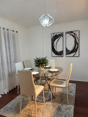 Un coin repas comprenant une table ronde en verre, des chaises blanches à coussin, un luminaire suspendu et deux tableaux abstraits accrochés au mur.