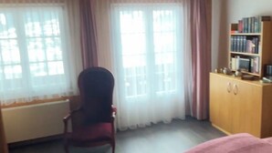 Een kamer met een houten plafond, een stoel, een bed, een raam met gordijnen en een boekenkast.
