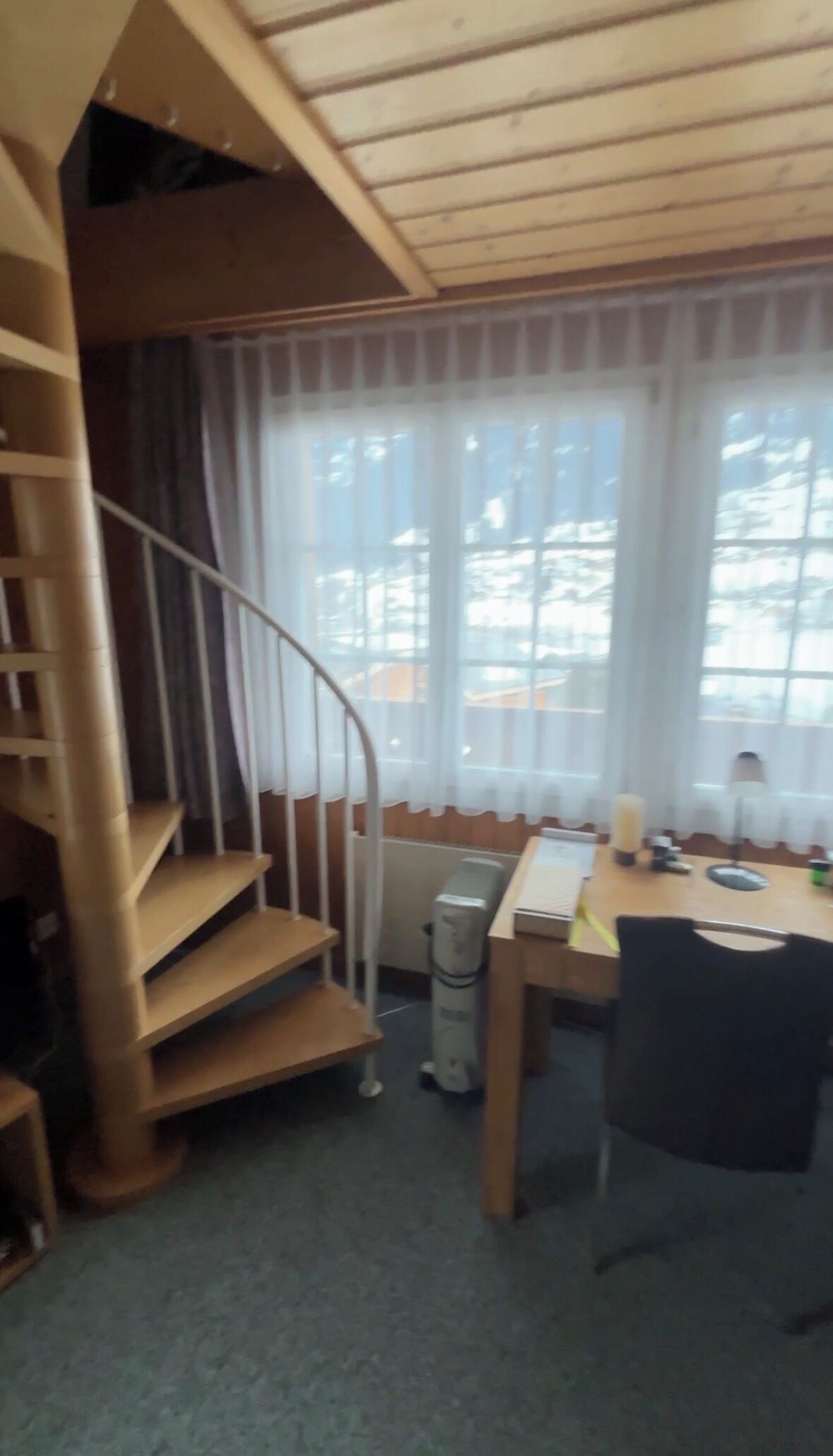 A room with a spiral staircase, a dining table with chairs, and a large window with curtains.