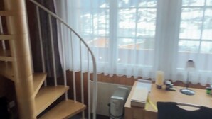 Een kamer met een spiltrap, een eettafel met stoelen en een groot raam met gordijnen.
