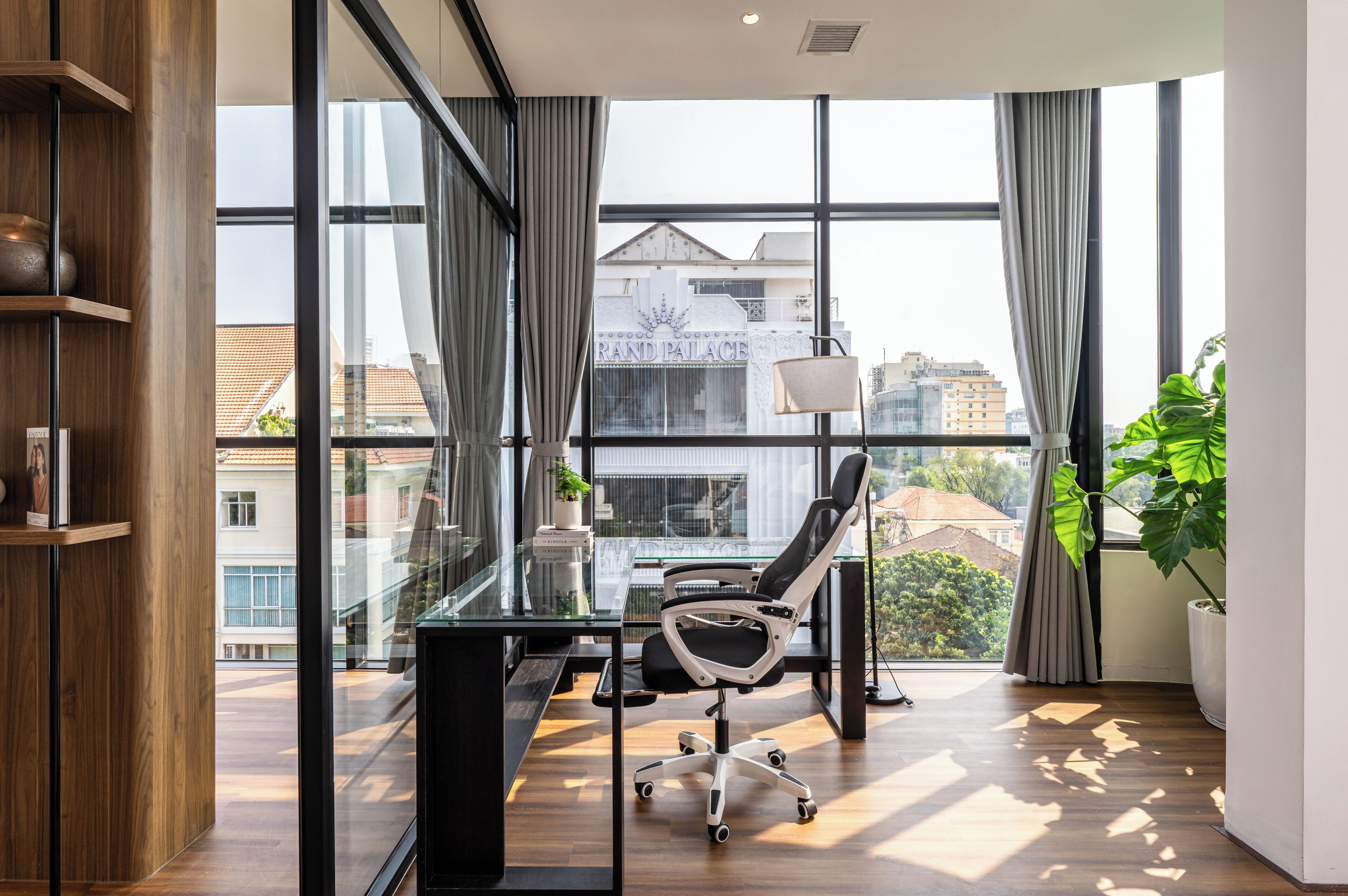 A modern office with a glass desk, a black office chair, a potted plant, and a view of a building labeled 'Grand Palace.'