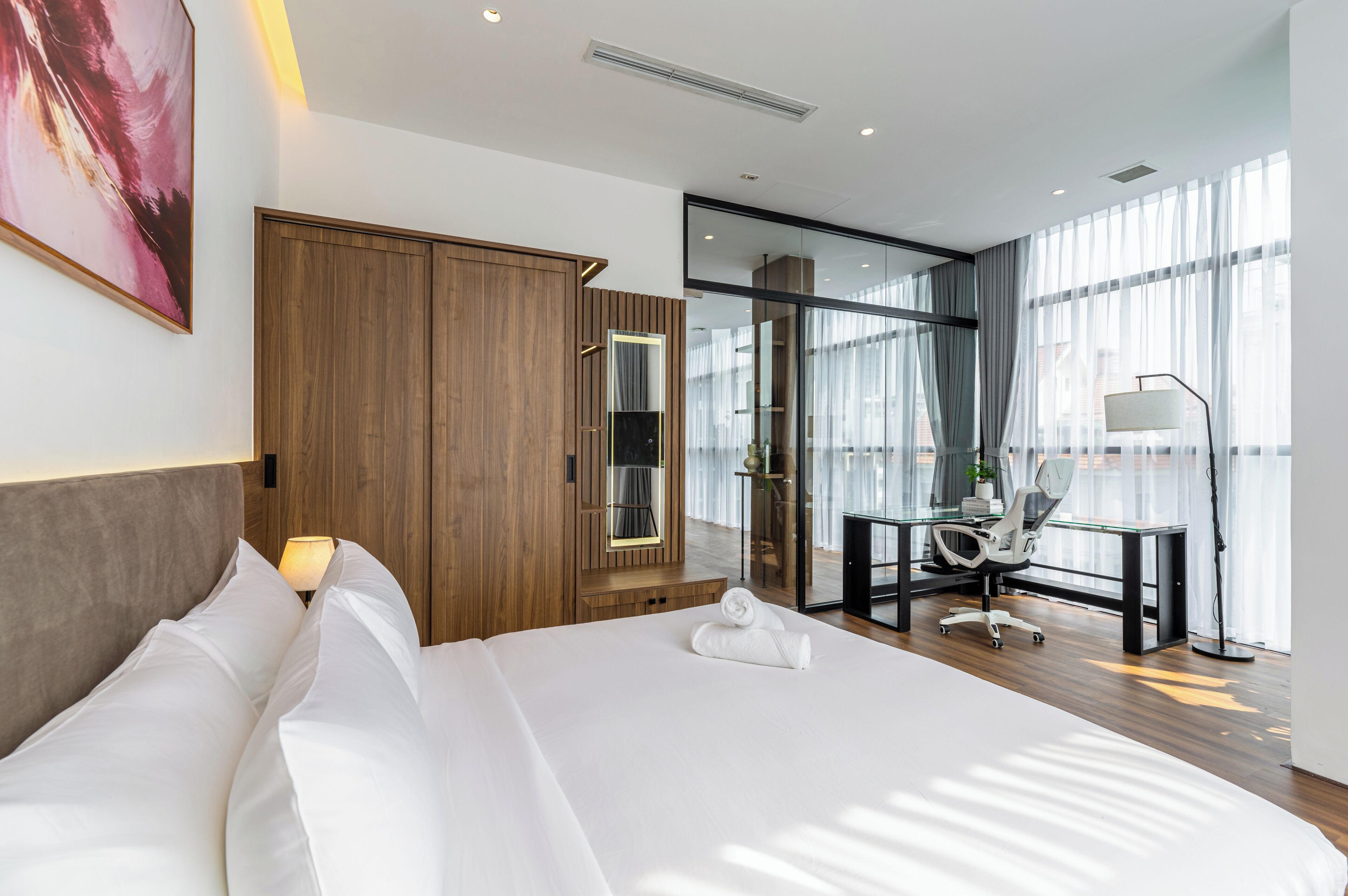 A modern hotel room with a large bed, a desk, a chair, and a glass door leading to a balcony.