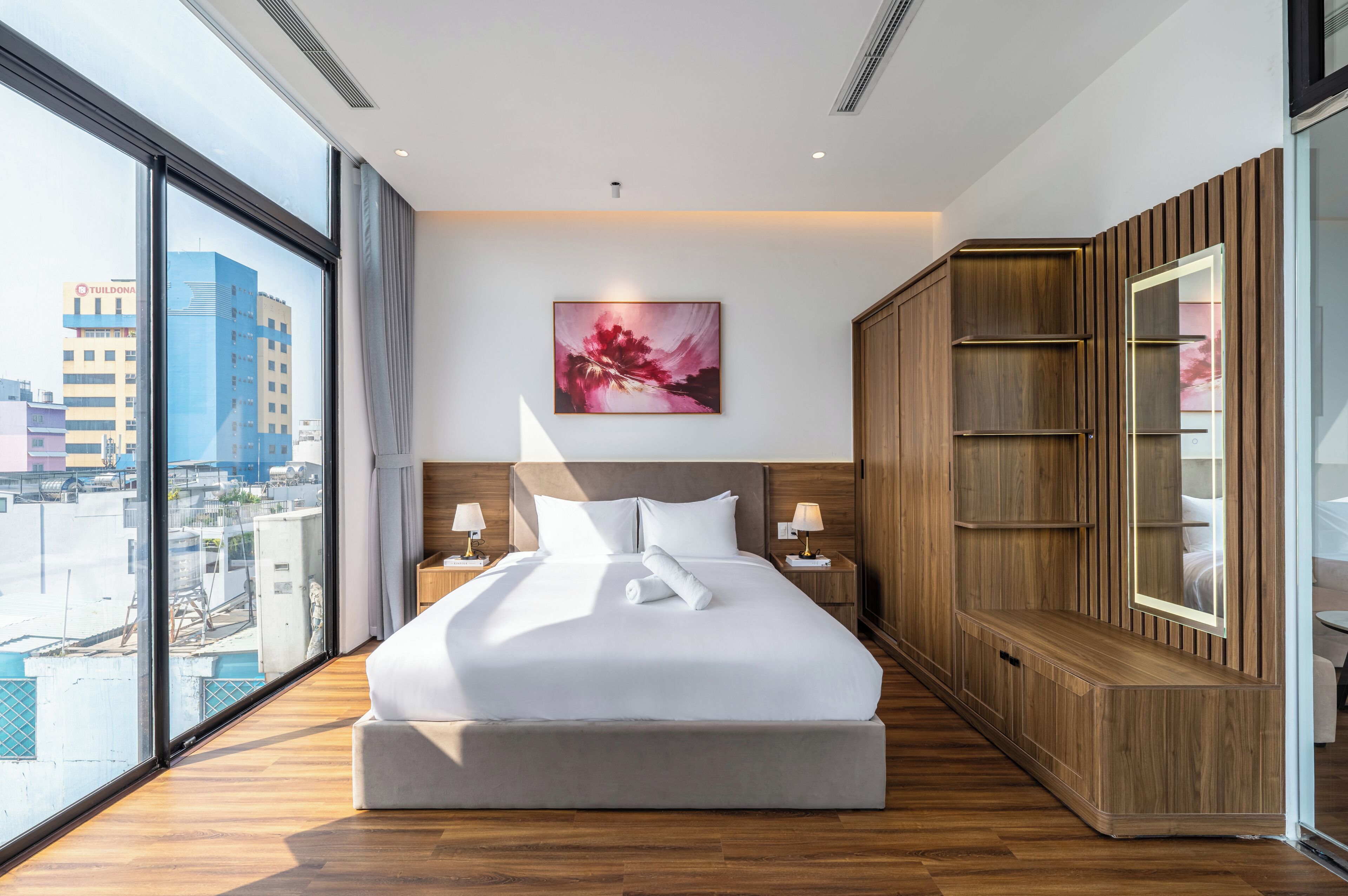 A hotel room with a large bed, wooden wardrobe, and a city view through floor-to-ceiling windows.