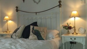 A bedroom with a white bed, decorative pillows, a headboard with a heart-shaped decoration, bedside tables with lamps, and a wall-mounted clock. - Cosy Romantic Cottage with Parking a short drive from the North Lakes & Pennines (Penrith)