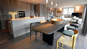 Una cocina moderna con una isla central, comedor con una mesa grande y sillas, y zona de estar con sofá y televisor.