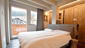Un dormitorio con una cama, un armario de madera, un taburete pequeño y vistas de un edificio a través de una ventana.