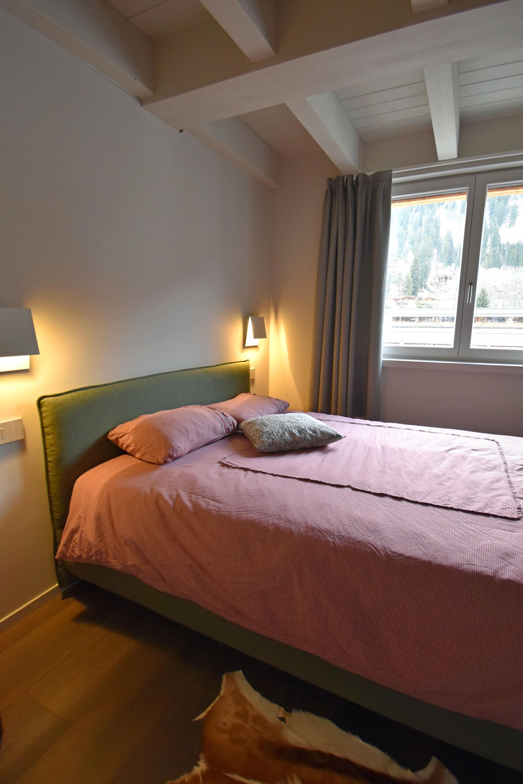 A bedroom with a bed, two wall-mounted lamps, a window with curtains, and a view of trees outside.