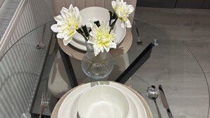 A glass table with white plates, a clear glass vase with white flowers, and a fork, knife, and spoon set. - Stylish 1 Bedroom Apartment in East Croydon - Fast Link To Gatwick Airport (Croydon)