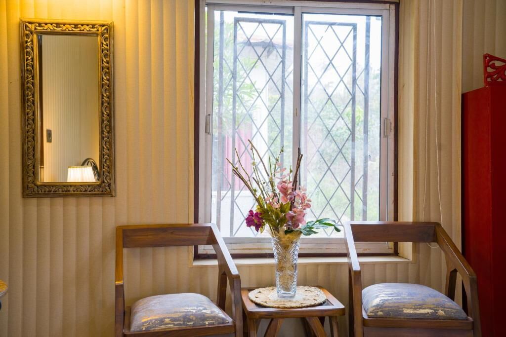 A room with a large window, a mirror, two wooden chairs, and a vase with flowers on a small table.
