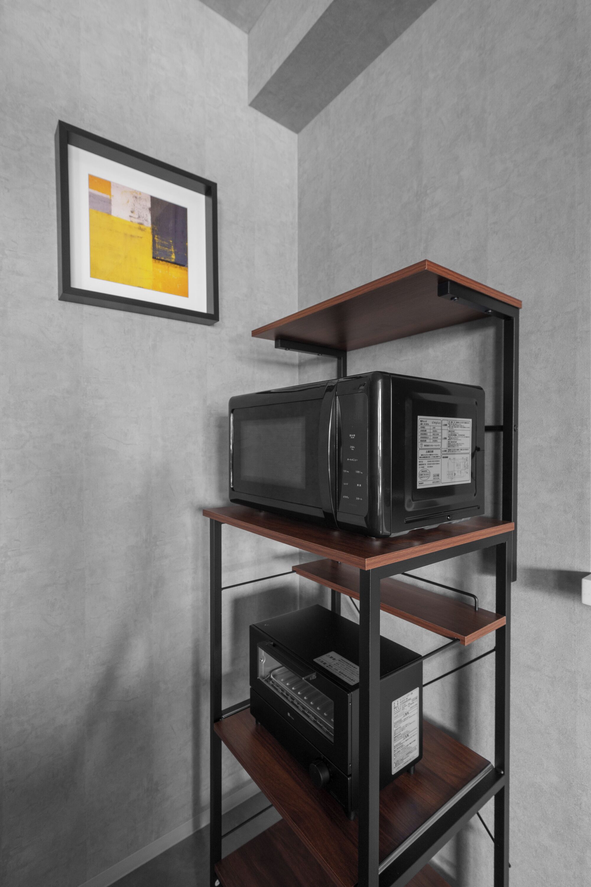 A black microwave on a wooden shelf unit against a grey wall, with a framed picture above it.