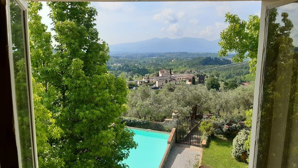 Une vue depuis une fenêtre donnant sur une piscine, une vég étation luxuriante et un village situé au loin.