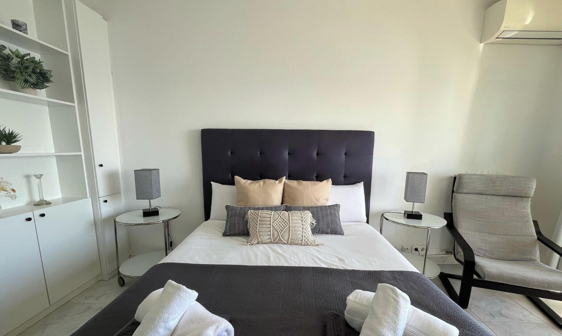 Une chambre d’hôtel moderne avec un grand lit, deux tables de chevet, une chaise et une armoire blanche.