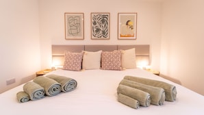 Ein ordentlich bezogenes Bett mit aufgerollten Handtüchern, ein Kopfteil mit drei gerahmten Kunstwerken und Nachttischlampen.
