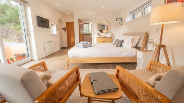 Ein Schlafzimmer mit einem großen Bett, einem Schreibtisch mit Laptop, Korbsesseln und Blick durch eine Glastür.