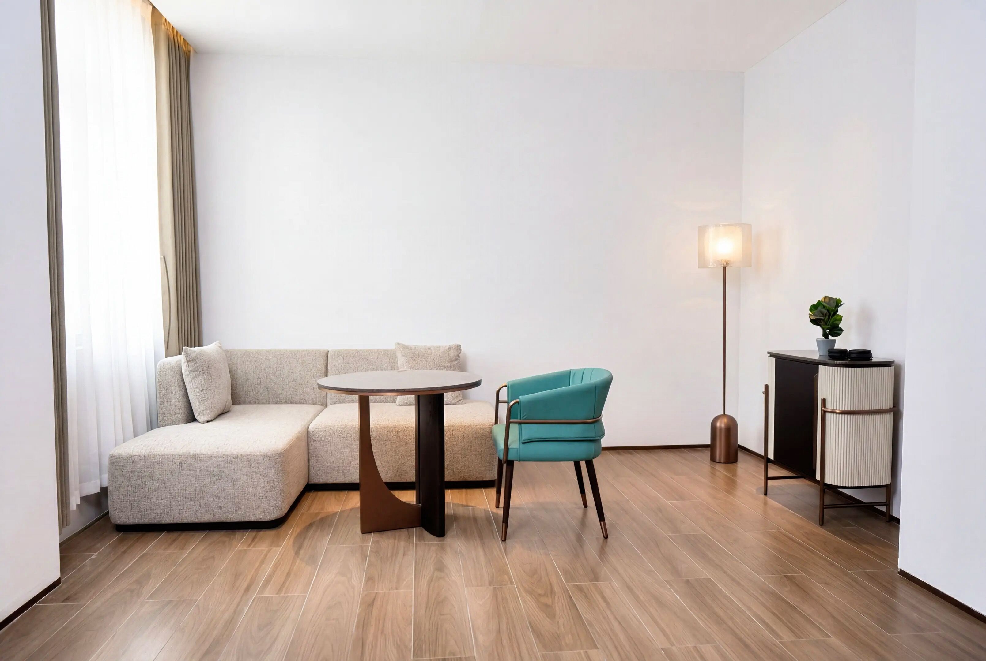 Una habitación de hotel moderna con un sofá seccional, una mesa redonda, una silla color esmeralda, una lámpara de pie y un mueble bajo.