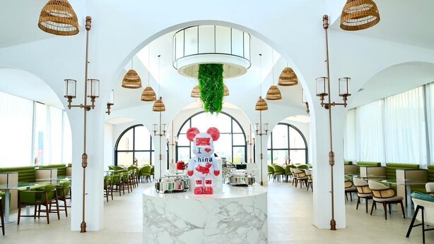 A spacious hotel lobby with a central reception desk, green seating, and a high ceiling with hanging lights.