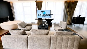 A modern hotel room with a large sofa set, a flat-screen TV, and a large window with curtains.