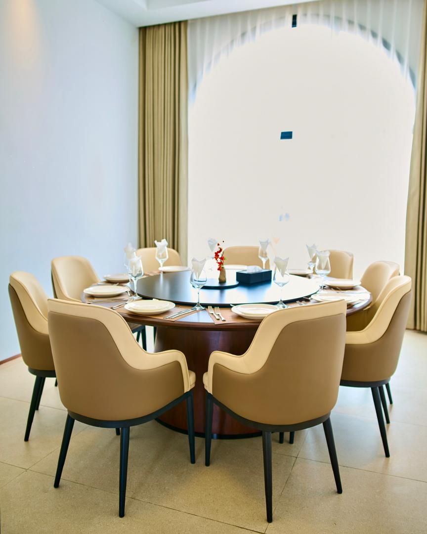 Een ronde eettafel met zes stoelen, elk met een donker houten onderstel en lichtgekleurde bekleding.