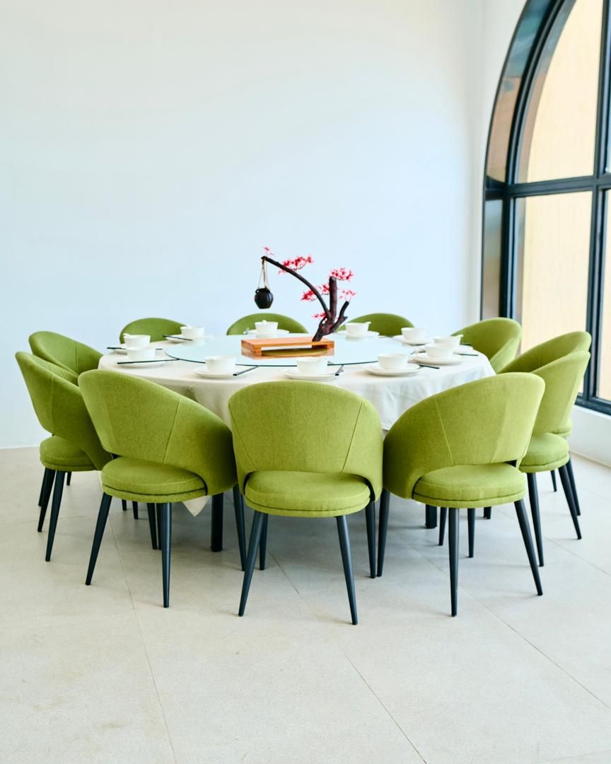 A round dining table with six green chairs, a white tablecloth, and a vase with pink flowers.