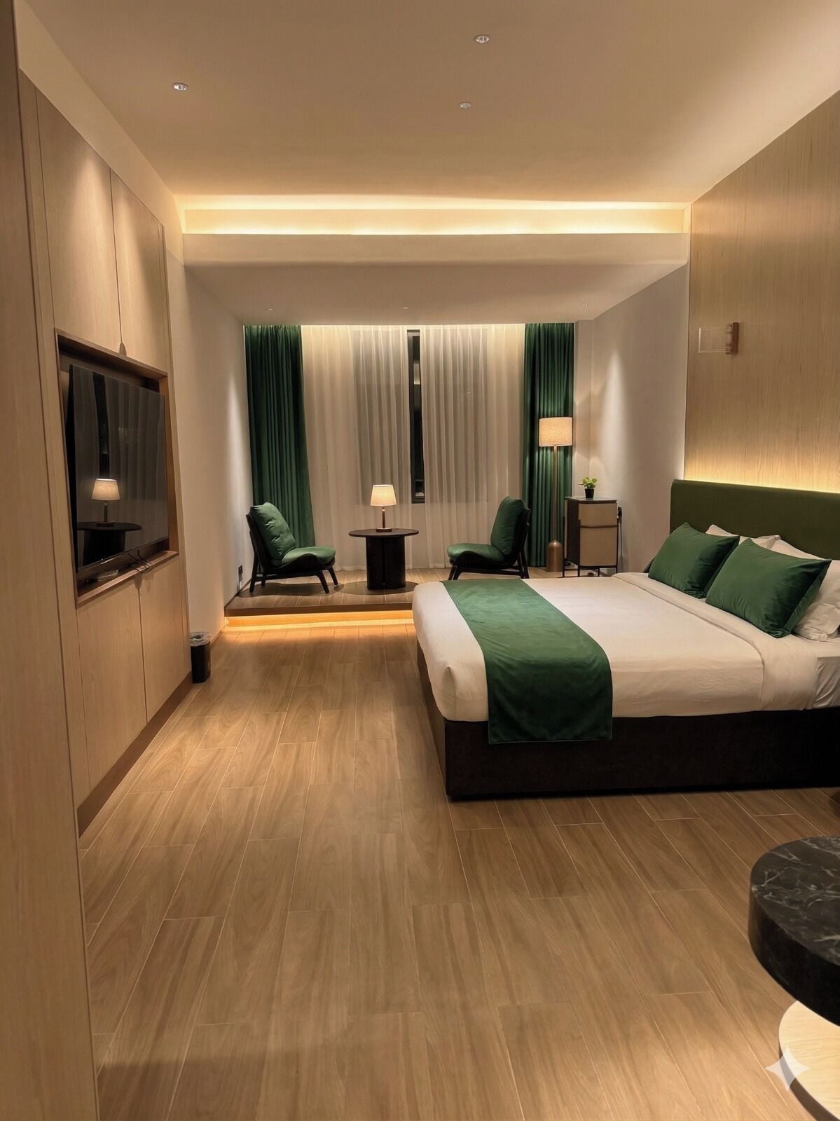 A modern hotel room with a large bed, two chairs, a small table, a TV, and a window with curtains.