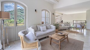 Ett rymligt vardagsrum med en soffa, fåtölj och soffbord, stora fönster med utsikt över grönområden och en balkong med utemöbler.