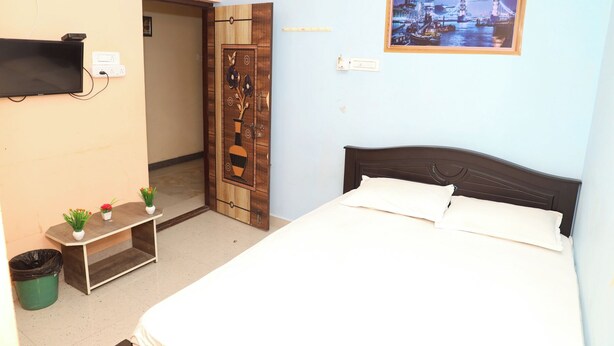 A bedroom with a bed, a TV mounted on the wall, a wooden door with a vase design, and a framed picture of a cityscape. - Akshaya Mahal A/C Rooms (Anapirandan)